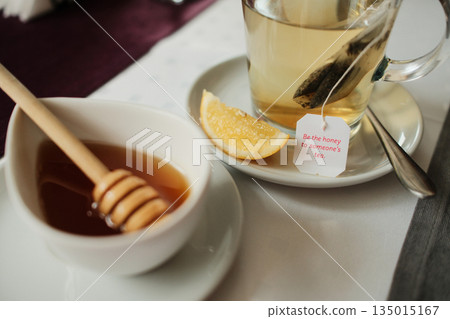 Warm herbal tea served with honey dipper and fresh lemon slice on a table. Calm lifestyle concept highlighting wellness, relaxation and slow moments in a cozy interior 135015167
