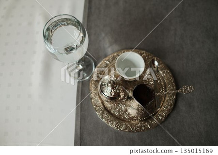 Bosnian coffee set with metal cezve, cup and water glass on a table. Concept of traditional serving, balance and hospitality Bosnian coffee set with metal cezve, cup and water glass on a table. Concept of traditional serving, balance and hospitality 135015169