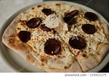 Fresh flatbread with melted cheese, sliced sausage and soft cheese topping served on a plate. Cozy Balkan cuisine concept focused on comfort food, simple ingredients and traditional dining atmosphere Fresh flatbread with melted cheese, sliced sausage and soft cheese topping served on a plate. Cozy Balkan cuisine concept focused on comfort food, simple ingredients and traditional dining atmosphere 135015175