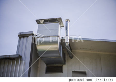Metal ventilation ducts extend from the roof of a building. Blue sky is present with no visible clouds. This scene shows part of a structure Metal ventilation ducts extend from the roof of a building. Blue sky is present with no visible clouds. This scene shows part of a structure 135015735