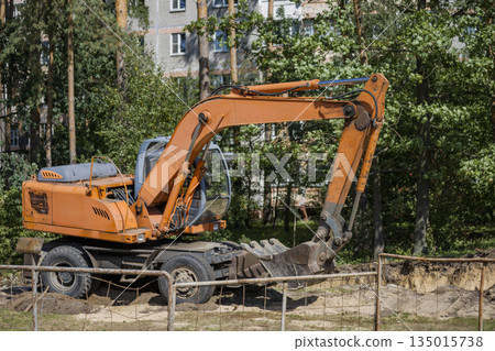 A construction vehicle works to dig a large hole in the ground by some buildings. Trees are visible in the background under clear weather conditions 135015738