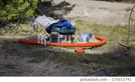 Tools, storage boxes, and red hoses are placed on the ground at a worksite. The area has some dirt and grass with scattered materials around Tools, storage boxes, and red hoses are placed on the ground at a worksite. The area has some dirt and grass with scattered materials around 135015741