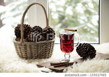 Cozy winter holiday scene with glass of hot mulled wine, cinnamon sticks and pine cones next to basket on windowsill with snowy trees outside the window. Cozy winter holiday scene with glass of hot mulled wine, cinnamon sticks and pine cones next to basket on windowsill with snowy trees outside the window. 135017485