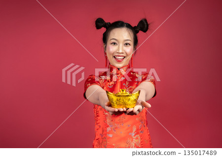 Portrait of smiling woman holding gold yuanbao toward camera on red background Portrait of smiling woman holding gold yuanbao toward camera on red background 135017489