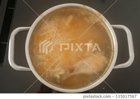 A pot of chicken soup simmering on the stove with noodles and vegetables visible in the broth. The warm, homemade dish conveys comfort, home cooking and the preparation of a simple traditional meal A pot of chicken soup simmering on the stove with noodles and vegetables visible in the broth. The warm, homemade dish conveys comfort, home cooking and the preparation of a simple traditional meal 135017567