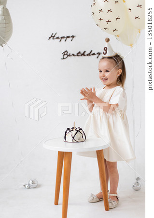 A young girl claps her hands with joy in front of a birthday cake, celebrating her third birthday with balloons and decorations. A young girl claps her hands with joy in front of a birthday cake, celebrating her third birthday with balloons and decorations. 135018255