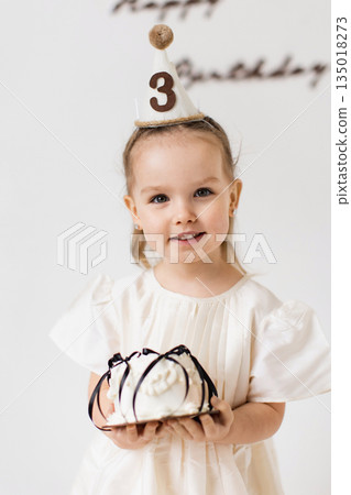 A young girl smiles while holding a birthday cake, wearing a party hat that indicates her age. A young girl smiles while holding a birthday cake, wearing a party hat that indicates her age. 135018273