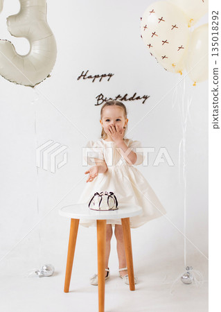 A young girl celebrates her birthday, standing in front of a white backdrop with balloons and a small cake. A young girl celebrates her birthday, standing in front of a white backdrop with balloons and a small cake. 135018292