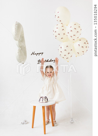 A young girl celebrates her birthday, reaching for the 'Happy Birthday' message with a cake and balloons. A young girl celebrates her birthday, reaching for the 'Happy Birthday' message with a cake and balloons. 135018294