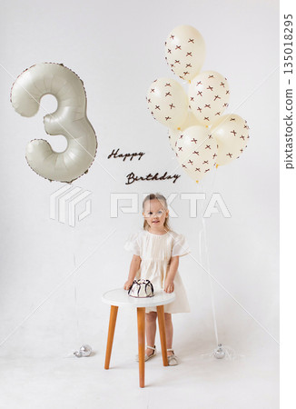 A young girl celebrates her third birthday with a cake and balloons in a bright, white setting. A young girl celebrates her third birthday with a cake and balloons in a bright, white setting. 135018295