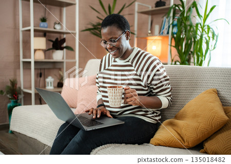 African American woman working from home, enjoying coffee on sofa African American woman working from home, enjoying coffee on sofa 135018482