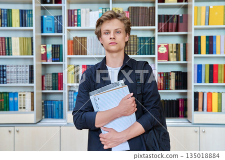 Portrait of serious teenage student with backpack textbooks in library 135018884