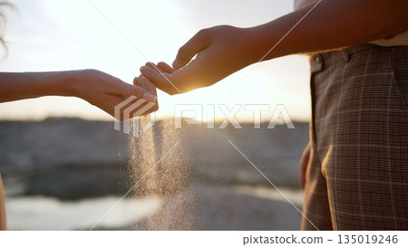 The hands of a young girl and a guy pour sand from the beach at sunset. Sea sand in a man's hand. man's hand holds sand. Sand falls out of your hands. Male and female hands pour sand. 135019246