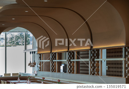 The dining space is a sophisticated area defined by graceful, sweeping arches and a palette of warm, earthy tones. Beneath the rhythmic curves of the ceiling, intimate tables are set with delicate 135019571