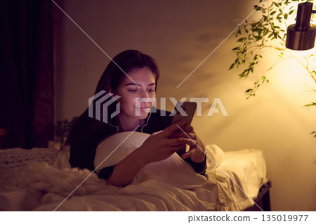 Woman using smartphone in bed under warm evening light. 135019977