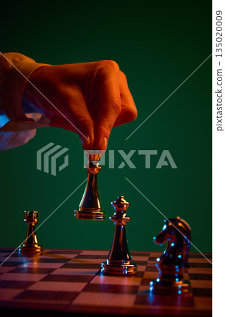 Hand holding queen above rival chess pieces under dramatic light. 135020009