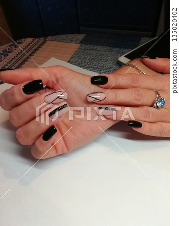 Manicured Hands Display. Hands showcasing manicured nails with black and nude polish, geometric patterns, rhinestones and glossy finish over white surface. Manicured Hands Display. Hands showcasing manicured nails with black and nude polish, geometric patterns, rhinestones and glossy finish over white surface. 135020402