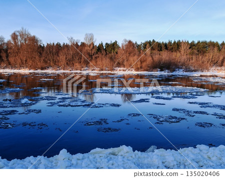 Frozen River with Floating Ice in Winter Landscape. Calm winter river partially covered with floating ice chunks reflecting trees and sky on cold sunny day. Frozen River with Floating Ice in Winter Landscape. Calm winter river partially covered with floating ice chunks reflecting trees and sky on cold sunny day. 135020406