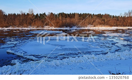 Winter River with Drifting Ice Floes at Sunset. Serene winter river scene with numerous floating ice chunks on water surface reflecting warm sunset light against forested shore. 135020768