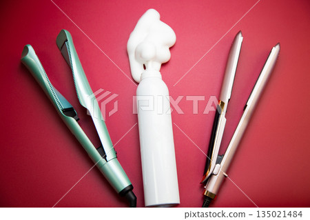 In the center are a white bottle of styling foam, around is a straightener and a curling iron to create hair curls on a pink background. The concept of hair care, creating a hairstyle and hair volume 135021484
