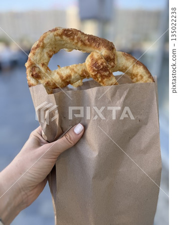 Hand holding fresh pretzel in paper bag outdoors on city street background 135022238