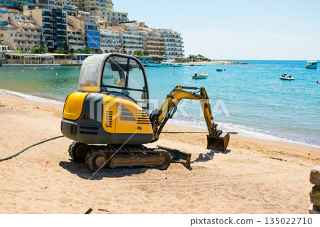 Compact excavator positioned on sandy beach near coastal buildings and calm sea. Urban development, tourism infrastructure, and transformation of natural shoreline environments. 135022710
