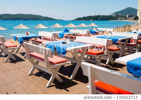 Rows of beachside cafe tables arranged along the shoreline in Montenegro. Order, repetition, and relaxed hospitality atmosphere before peak seaside dining hours. Rows of beachside cafe tables arranged along the shoreline in Montenegro. Order, repetition, and relaxed hospitality atmosphere before peak seaside dining hours. 135022713