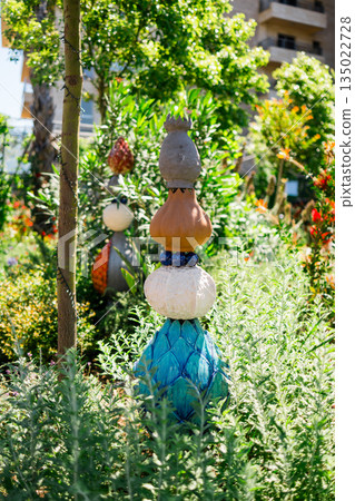 Decorative garden sculpture surrounded by lush green plants in sunlight. Artistic outdoor installation, creative landscape design and serene residential environment blending art with nature 135022728