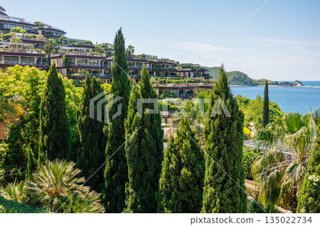 Modern resort buildings surrounded by lush greenery near the sea. Mediterranean Montenegro coastal lifestyle, luxury hospitality, summer vacation atmosphere and architectural harmony with natural Modern resort buildings surrounded by lush greenery near the sea. Mediterranean Montenegro coastal lifestyle, luxury hospitality, summer vacation atmosphere and architectural harmony with natural 135022734