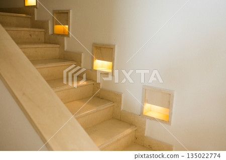 Interior staircase with integrated wall lighting and stone steps. Safe movement design, architectural guidance, functional illumination and calm residential circulation space Interior staircase with integrated wall lighting and stone steps. Safe movement design, architectural guidance, functional illumination and calm residential circulation space 135022774