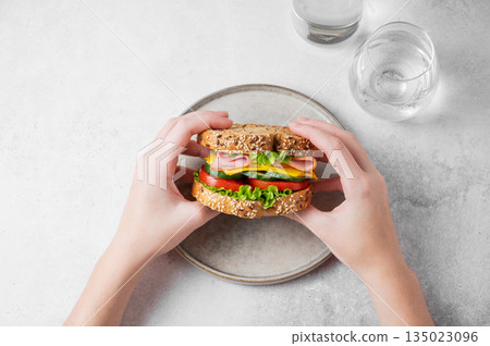 A hands holding a sandwich with ham, cheese, lettuce, and vegetable over a plate  135023096