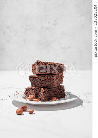 Chocolate brownie on a plate on a marble background with nuts and morning shadow. Chocolate brownie on a plate on a marble background with nuts and morning shadow. 135023104