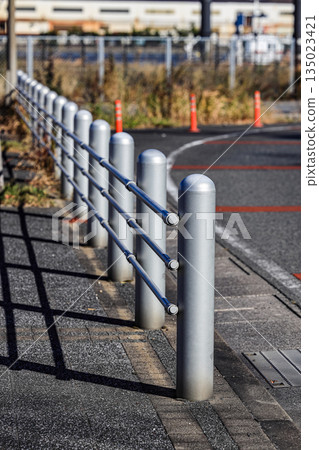 Row of metal safety bollards installed along the roadside near a modern residential area. The bollards act as vehicle barriers for traffic control and pedestrian safety. Row of metal safety bollards installed along the roadside near a modern residential area. The bollards act as vehicle barriers for traffic control and pedestrian safety. 135023421