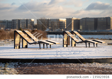 Empty recreational seats covered in snow standing in city park near river facing bay on winter day. 135023916