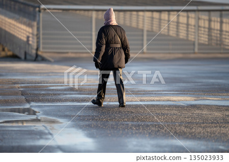 Woman walking on frozen asphalt sidewalk with technical salt and substances sprinkled over surface. 135023933