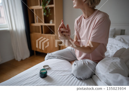 Middle aged woman doing beauty routine rituals in morning, applying hand cream from jar on skin. 135023934