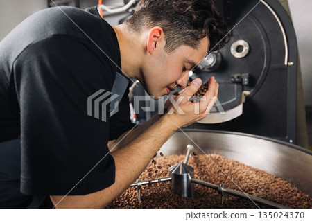 Side view of young worker smelling fresh roasted coffee beans above the machine drum Side view of young worker smelling fresh roasted coffee beans above the machine drum 135024370