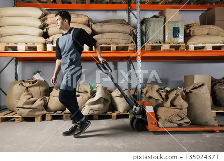 Male in apron pulling weighing pallet truck with coffee bags. Warehouse worker moving hydraulic pallet truck. 135024371
