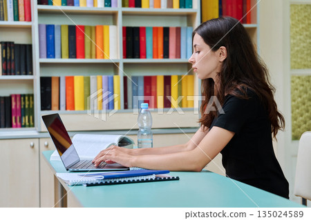Teenage high school student girl studies in the library with laptop and books Teenage high school student girl studies in the library with laptop and books 135024589