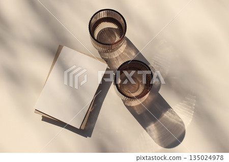 Blank greeting card, invitation mockup on beige table background. Two colored water glasses, bathed in sunlight casting shadows of leaves. Summer stationery, refreshing concept. Flat lay, top view 135024978