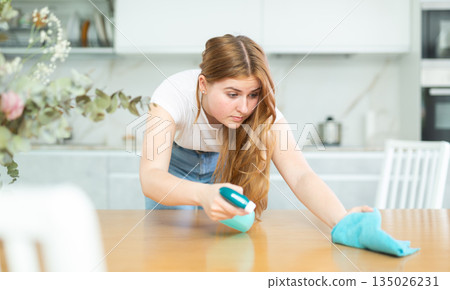 Attentive European teen girl in casual clothes spraying detergent on microfiber rag and wiping countertop in cozy cuisine 135026231
