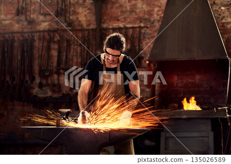 Blacksmith, man and metal workshop and sparks, manual forge working on hot steel with hammer, and fire. Worker, person welding or iron tools manufacturing and expert, trade and dark workspace Blacksmith, man and metal workshop and sparks, manual forge working on hot steel with hammer, and fire. Worker, person welding or iron tools manufacturing and expert, trade and dark workspace 135026589