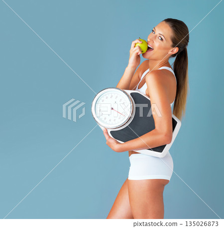 Woman is eating apple, fruit and scale with diet and health for weight loss in portrait isolated on blue background. Healthy food, female person and organic in studio with wellness and mockup space Woman is eating apple, fruit and scale with diet and health for weight loss in portrait isolated on blue background. Healthy food, female person and organic in studio with wellness and mockup space 135026873
