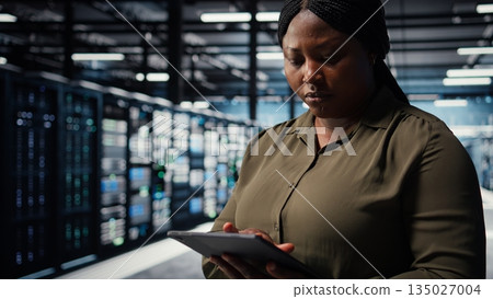 Data center software developer uses tablet to develop disaster recovery plans, minimizing downtime. IT worker in server hub enhancing data protection protocols using device, camera A close up Data center software developer uses tablet to develop disaster recovery plans, minimizing downtime. IT worker in server hub enhancing data protection protocols using device, camera A close up 135027004