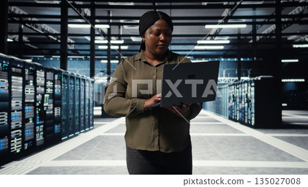 Supervisor in data center using laptop to coordinate deployment of new server architecture. Server farm IT specialist using notebook to do rollout of cybersecurity framework, protecting data, camera A Supervisor in data center using laptop to coordinate deployment of new server architecture. Server farm IT specialist using notebook to do rollout of cybersecurity framework, protecting data, camera A 135027008