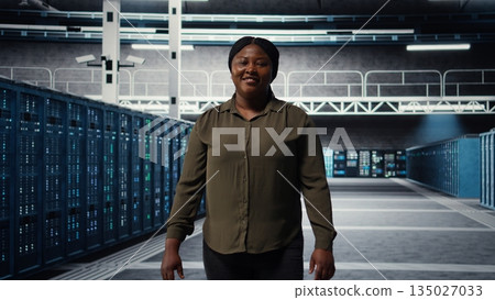 Portrait of smiling admin walking in data center, overseeing large scale IT infrastructure to maintain seamless operations. Happy server farm employee pleased by increased uptime, camera A Portrait of smiling admin walking in data center, overseeing large scale IT infrastructure to maintain seamless operations. Happy server farm employee pleased by increased uptime, camera A 135027033