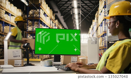 Female african american worker using pc with isolated mockup display, wearing hi vis vest and helmet in warehouse environment. E-commerce retail operations surrounded by big boxes. Camera A. Female african american worker using pc with isolated mockup display, wearing hi vis vest and helmet in warehouse environment. E-commerce retail operations surrounded by big boxes. Camera A. 135027048