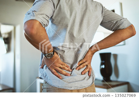 Hands, back pain and injury with a man standing in the living room of his home, holding his spine in discomfort. Health, medical and anatomy with a male person in a house feeling a muscle cramp Hands, back pain and injury with a man standing in the living room of his home, holding his spine in discomfort. Health, medical and anatomy with a male person in a house feeling a muscle cramp 135027069
