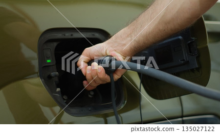 Close up shot of driver recharging EV vehicle battery powering green tech electric motor. Person recharges battery of EV car by plugging into renewable energy power source Close up shot of driver recharging EV vehicle battery powering green tech electric motor. Person recharges battery of EV car by plugging into renewable energy power source 135027122