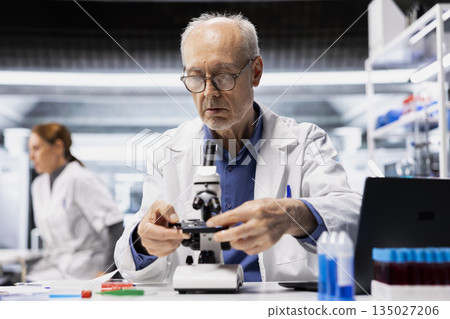 Senior lab manager checks product surface for tiny defects using microscope. Elderly research facility professional ensures manufacturing standards, examines small parts with optical device Senior lab manager checks product surface for tiny defects using microscope. Elderly research facility professional ensures manufacturing standards, examines small parts with optical device 135027206